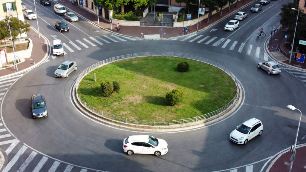 Rotatoria vista dall’alto con auto in circolazione, un’auto in attesa all’ingresso e un ciclista superato a distanza, per l’articolo sulle novità del Codice della strada