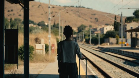 Giovane donna con valigia di spalle su un binario di una stazione siciliana, sullo sfondo un paese dell’entroterra