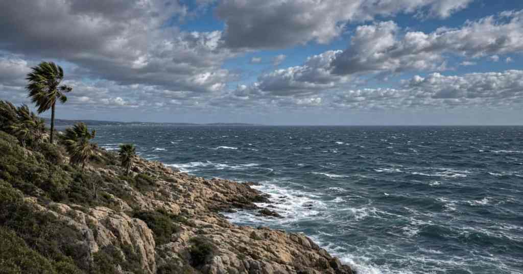 Illustrazione del meteo weekend Sicilia: costa mediterranea con nubi in transito e mare mosso dal vento (immagine generata con IA, a scopo illustrativo)